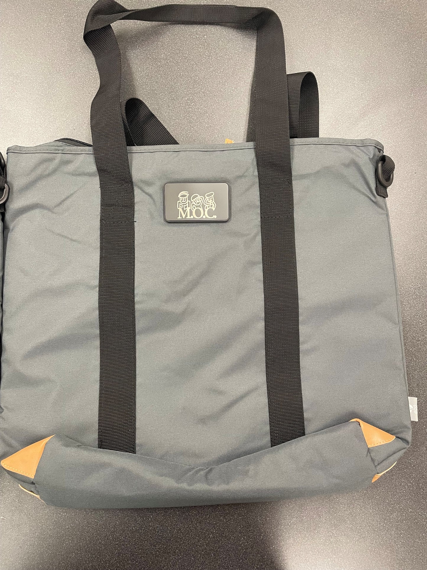 A gray fabric tote bag with black shoulder straps and a small rectangular logo patch reading 'MOC' on the front, photographed on a gray surface.