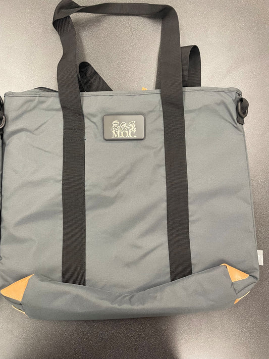 A gray fabric tote bag with black shoulder straps and a small rectangular logo patch reading 'MOC' on the front, photographed on a gray surface.